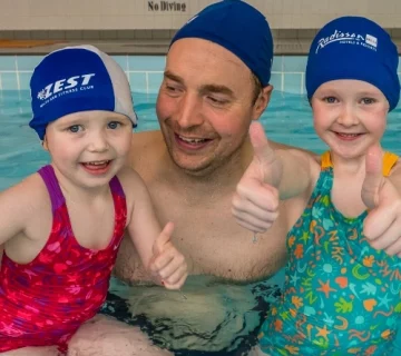 A smiling adult, wearing a blue swim cap, stands in a swimming pool with two young children. Both children are also smiling, wearing swim caps and swimsuits—one in a red swimsuit and the other in a green one with colorful patterns. They are giving a thumbs up.