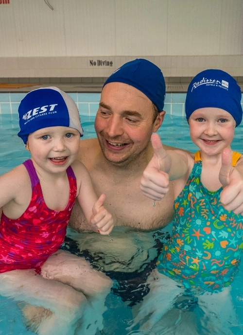 A smiling adult, wearing a blue swim cap, stands in a swimming pool with two young children. Both children are also smiling, wearing swim caps and swimsuits—one in a red swimsuit and the other in a green one with colorful patterns. They are giving a thumbs up.