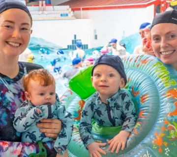 Two adults and two young children dressed in swimwear pose at an indoor swimming pool. One adult holds a baby, and the other stands beside a child who is sitting in a green inflatable trainer seat. The pool is bustling with other swimmers in the background.