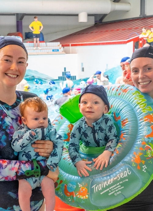 Two adults and two young children dressed in swimwear pose at an indoor swimming pool. One adult holds a baby, and the other stands beside a child who is sitting in a green inflatable trainer seat. The pool is bustling with other swimmers in the background.