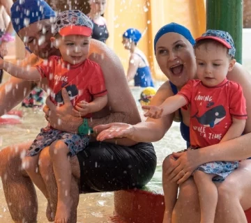 A family sits by a water play area, playfully splashing around. The father and mother, both wearing blue swim caps, each hold a young child in matching red shirts and swim caps. Water droplets surround them as they smile and enjoy the playful moment.