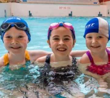 Three children smiling and standing in the shallow end of an indoor swimming pool. They are wearing swim caps and goggles. The background shows "Wave Pool" written on the wall, with a sign indicating the deep end at 1.5 meters.