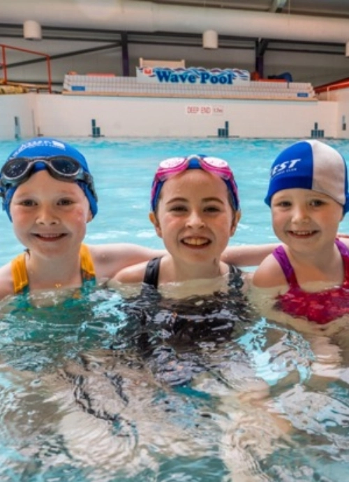 Three children smiling and standing in the shallow end of an indoor swimming pool. They are wearing swim caps and goggles. The background shows "Wave Pool" written on the wall, with a sign indicating the deep end at 1.5 meters.