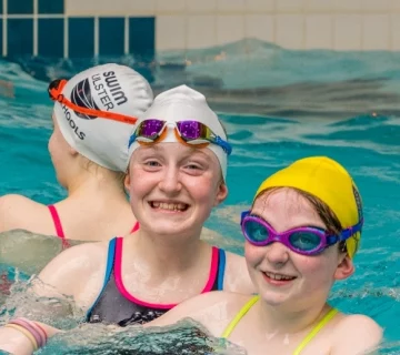 Three children, wearing swimming caps and goggles, smile as they swim in a pool. The child in the middle has a white cap and is grinning widely, while the child on the right has a yellow cap and pink goggles. The child on the left faces away from the camera.