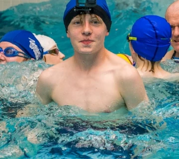 A young swimmer wearing a swim cap and a mounted camera on their head stands chest-deep in a pool, surrounded by other swimmers. The water splashes around them, and swimming caps and goggles are visible on other swimmers in the background.