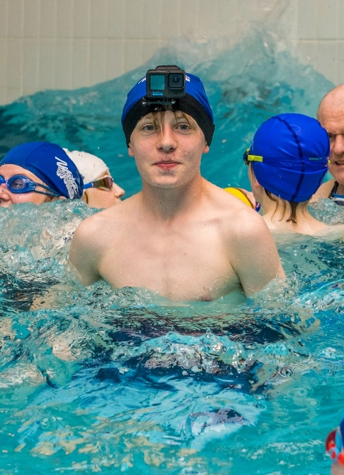 A young swimmer wearing a swim cap and a mounted camera on their head stands chest-deep in a pool, surrounded by other swimmers. The water splashes around them, and swimming caps and goggles are visible on other swimmers in the background.