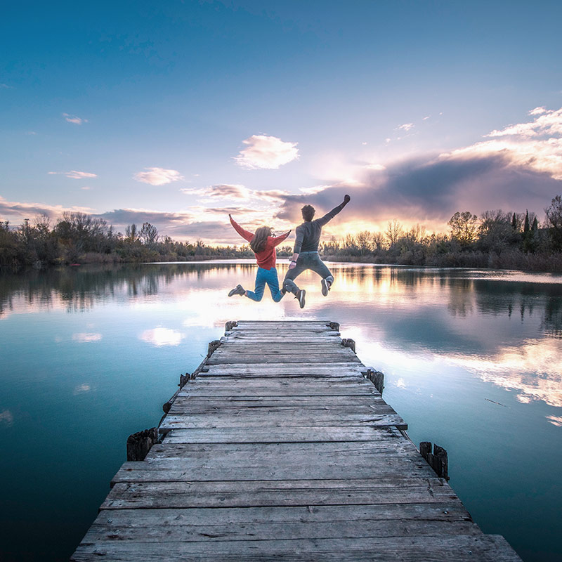 Two people are jumping joyfully at the end of a wooden dock that extends into a calm lake. The sky above is a vibrant mix of blues and pinks with scattered clouds, reflecting beautifully in the water. Trees and shrubs line the distant shore.