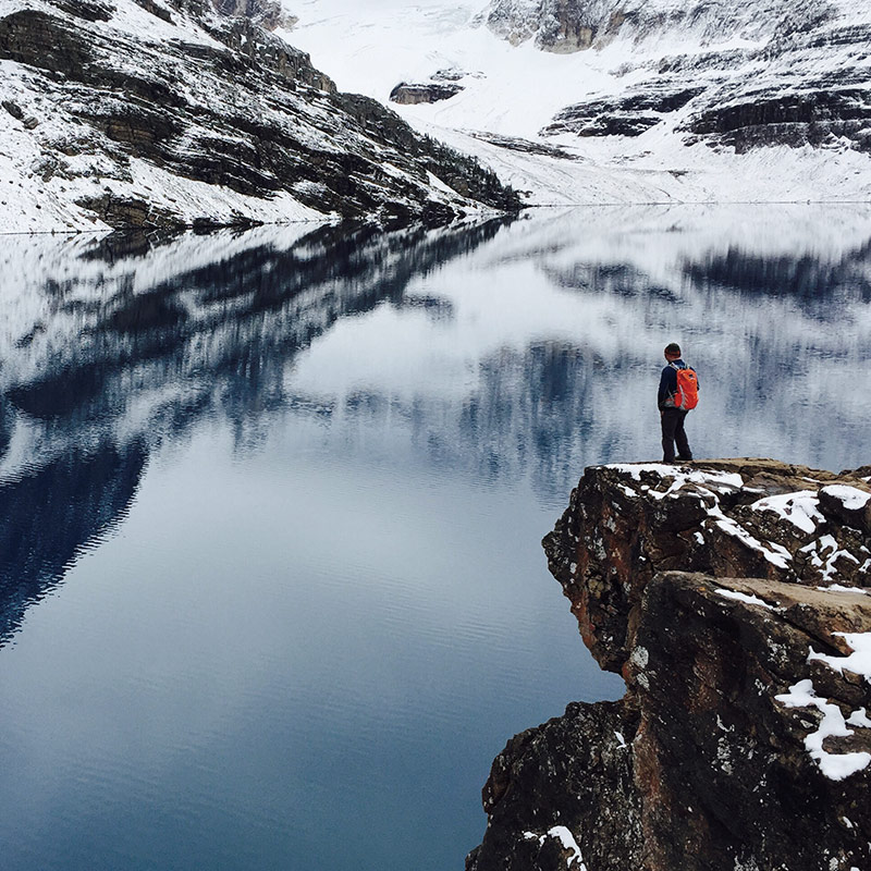 A person wearing a red backpack stands on a rocky ledge overlooking a calm, reflective lake surrounded by snow-covered mountains. The serene landscape captures the tranquility of the winter scenery.