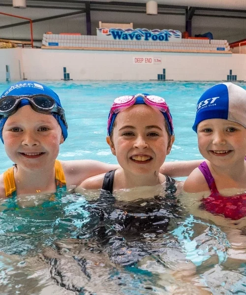 Three children smiling in a swimming pool. They are wearing swim caps and goggles. The background shows a large wave pool with a sign for "Wave Pool" and a "Deep End" marker. The kids are standing in the water with their arms around each other.