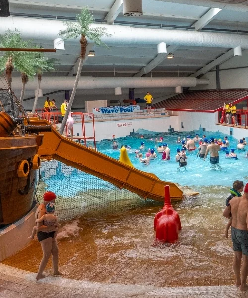 Indoor wave pool with numerous people enjoying the water. A red and yellow waterslide resembling a pirate ship enters the pool from the left. Lifeguards in yellow shirts are stationed around the pool. Fake palm trees and various pool toys are visible.