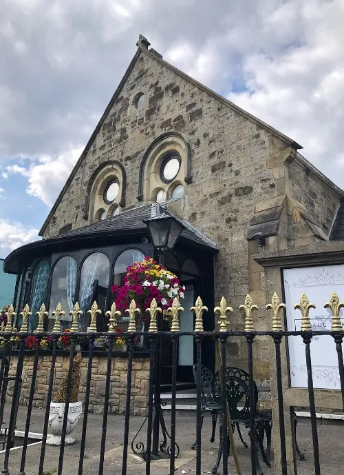 A stone building with a steeply pitched roof and circular windows, featuring a glass-enclosed porch filled with colorful flowers. A black wrought-iron fence with gold accents is in the foreground. The sky above has scattered clouds.