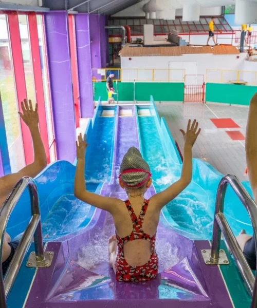 A child wearing a swim cap and a patterned swimsuit sits at the top of a waterslide with arms raised. The slide, a vibrant blue and purple, leads downward in an indoor water park. Bright, multicolored surroundings and another person are visible in the background.