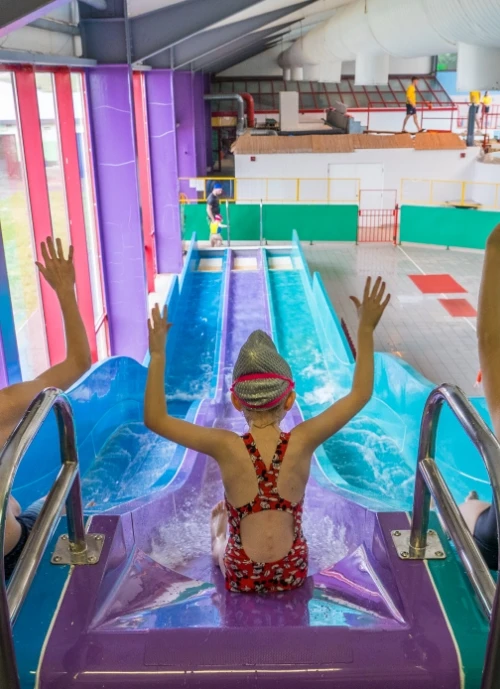 A child wearing a swim cap and a patterned swimsuit sits at the top of a waterslide with arms raised. The slide, a vibrant blue and purple, leads downward in an indoor water park. Bright, multicolored surroundings and another person are visible in the background.