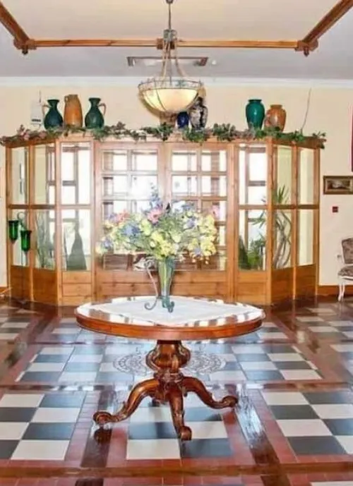 A well-lit interior features a wooden table with ornate legs, topped by a vase filled with colorful flowers. The floor is tiled in a geometric pattern, and a wooden glass-enclosed display with green vases, plants, and decorative items lines the back wall.