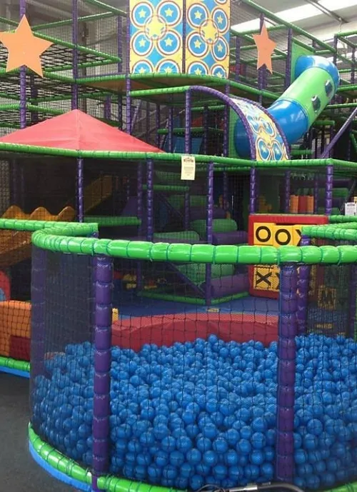 Indoor children's play area with a ball pit filled with blue balls, surrounded by a green and purple padded fence. The background features a large multi-level play structure with slides, climbing nets, and decorative patterns with stars.