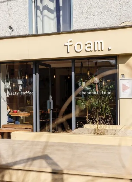 A cozy café named "foam." featuring large windows and outdoor seating. The signage on the windows reads "specialty coffee" and "seasonal food." Inside, a mix of wooden tables and lush greenery can be seen, offering a welcoming atmosphere.