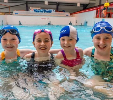 Four children in swimsuits and swim caps smile at the camera while standing in the shallow end of an indoor pool. A sign reading "Wave Pool" is visible in the background above the pool.