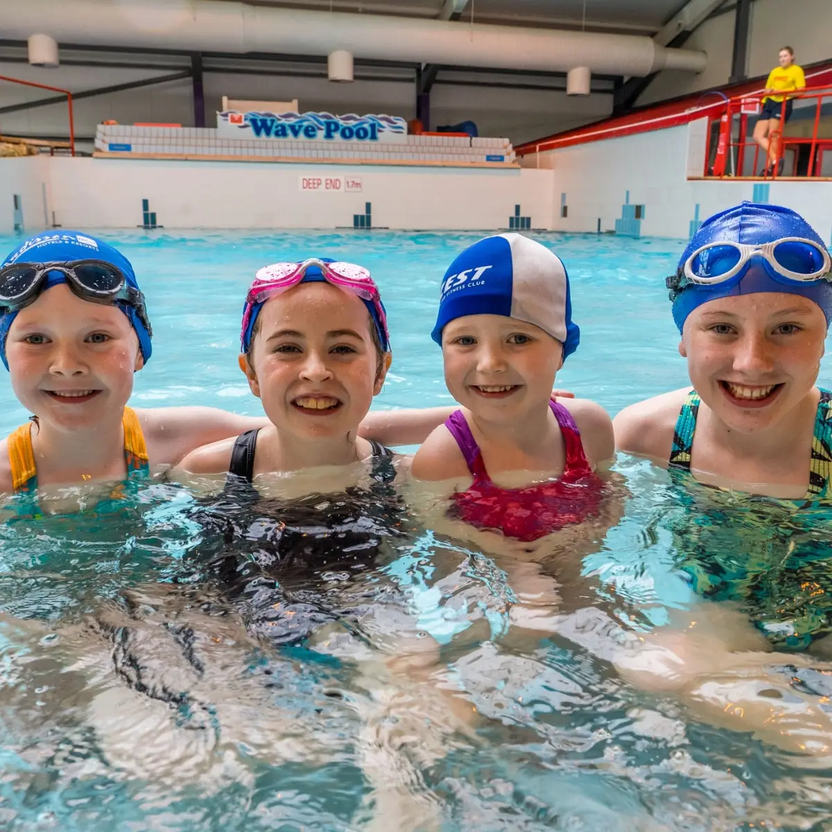 Four children in swimsuits and swim caps smile at the camera while standing in the shallow end of an indoor pool. A sign reading "Wave Pool" is visible in the background above the pool.