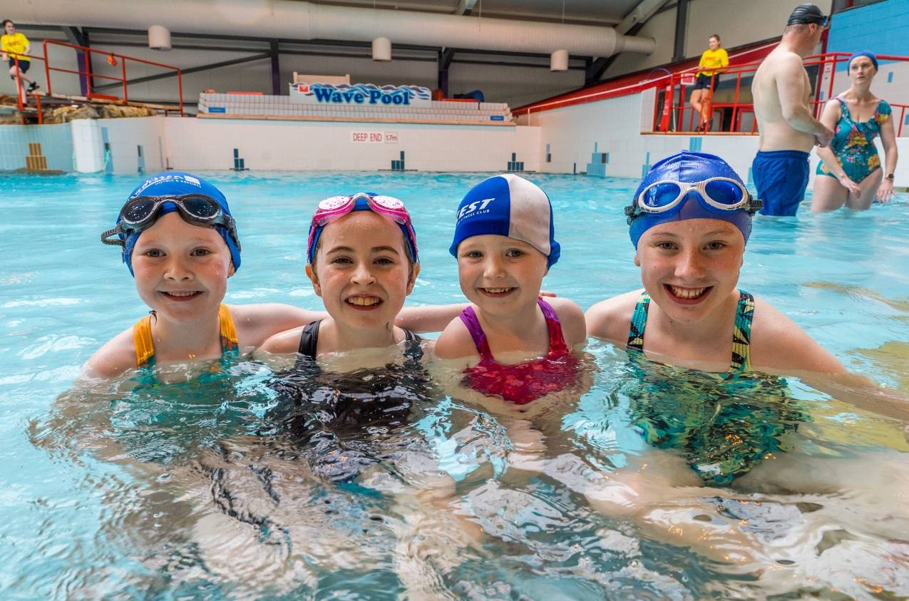 A group of four smiling girls wearing swim caps and goggles stand together in an indoor swimming pool. Behind them are other swimmers and the sign "Wave Pool" on the wall. The pool area is bright, with red railings and white tiles lining the pool.