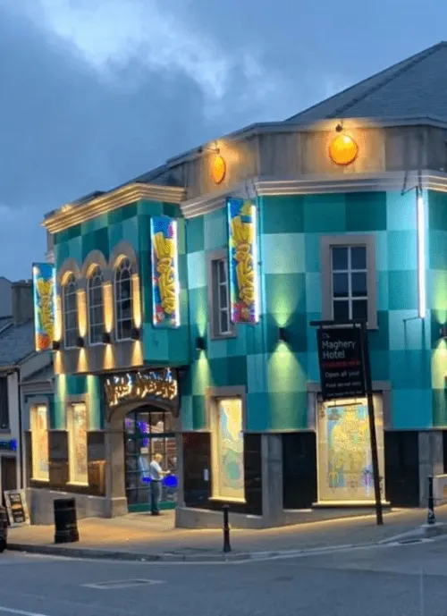 A brightly lit building with a checkered turquoise facade, colorful banners, and large windows. The sign reads "Maghery Hotel." The sky is overcast, and the street in front of the hotel is quiet with minimal traffic.