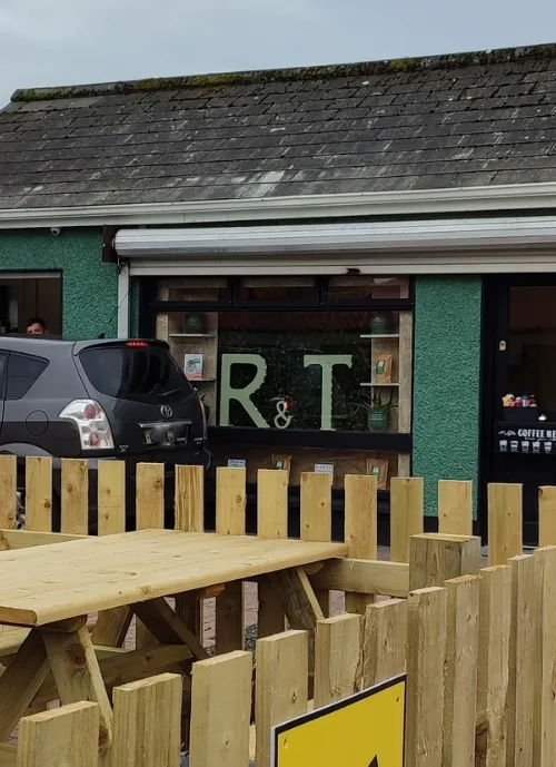 A photo of a cafe with a green exterior and a sign reading "R&T" in the window. A black SUV is parked nearby. There is a wooden fence and picnic tables in the foreground, and a yellow sign with a black arrow. The sky is overcast.