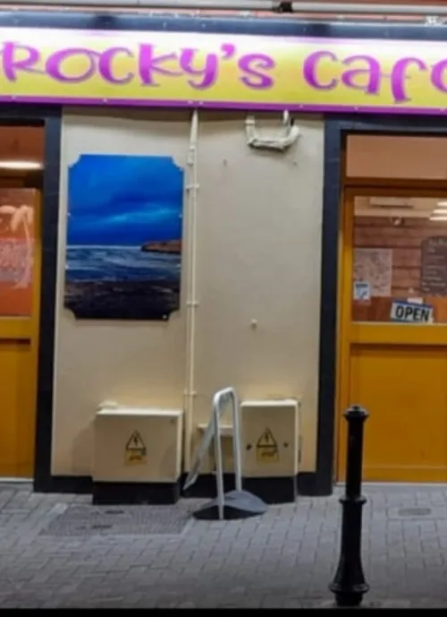 The entrance of "Rocky's Cafe" with a yellow sign. There's a painting of a coastal scene on the exterior wall, an "Open" sign in the window, two metal bins, and a bike rack on the sidewalk. The door and windows are framed in orange.