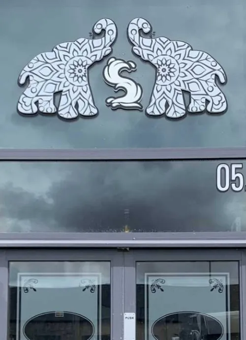 A glass door with an intricate design featuring two stylized elephants forming an arch with their trunks and a decorative "S" between them. Above the door is the number "05," and reflections of clouds can be seen on the glass.