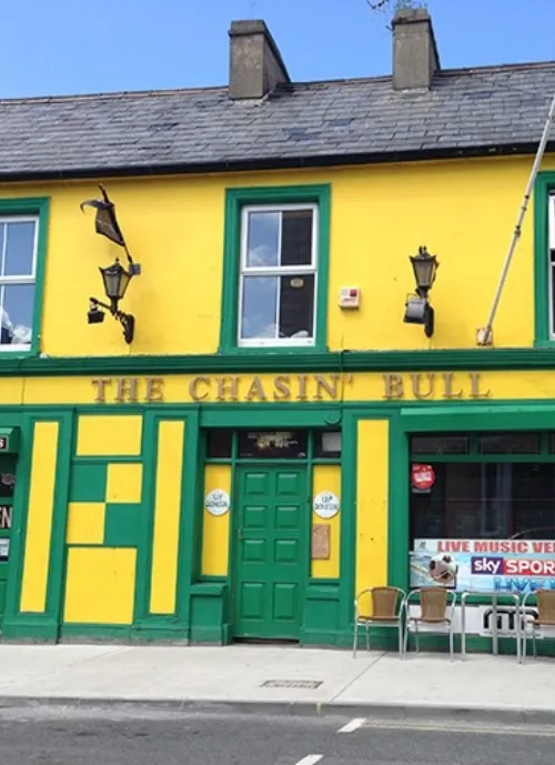 The image shows a bright yellow and green building that houses "The Chasin' Bull" pub. The pub has a few outdoor tables and chairs. A sign in the window advertises live music and sports on Sky Sports. The building has three windows and a central door.