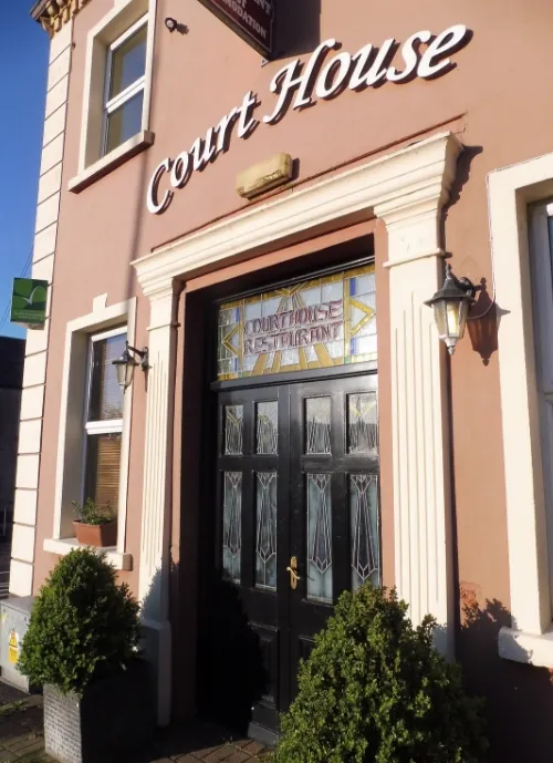 A brown building facade with a sign above the door reading "Court House." The entrance features a decorative glass panel above black double doors, flanked by two potted plants. To the left, there's another green sign, and sunlight casts shadows on the building.