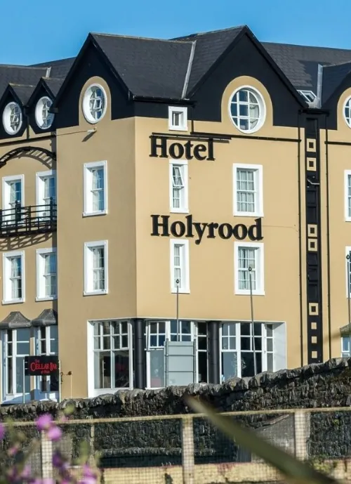 A multi-story building with beige walls and dark roof displaying the sign "Hotel Holyrood" in large letters on the facade. The architecture features several circular windows and a small balcony. Flowers and a stone wall are visible in the foreground.