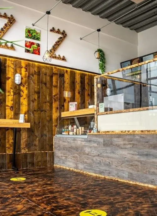 A modern café interior with rustic wooden walls and a counter. The counter features sandwiches and beverages. There are social distancing markers on the polished floor. The ceiling has exposed metal beams, and the wall decor includes photos of fresh produce.