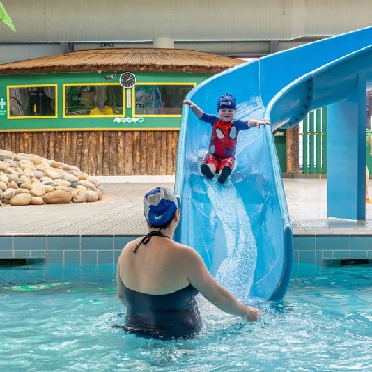 A baby wearing a blue and red swimsuit and a swim cap slides down a blue water slide into a shallow swimming pool, with an adult in a black swimsuit and swim cap waiting at the bottom. The background features a tropical-themed structure and stone decorations.