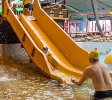 Children in swimwear are sliding down two yellow water slides at an indoor water park. An adult in a yellow swim cap is waiting at the bottom of the slides to catch them. The environment features a tropical theme with artificial palm trees and rocks.