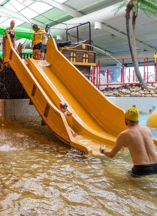 Children in swimwear are sliding down two yellow water slides at an indoor water park. An adult in a yellow swim cap is waiting at the bottom of the slides to catch them. The environment features a tropical theme with artificial palm trees and rocks.