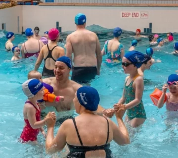 A crowded indoor wave pool with adults and children wearing swimsuits and swim caps. Some people are holding hands or playing in the water. A sign reads "Wave Pool" and "Deep End 1.7m" above the pool. The atmosphere is lively and engaging.