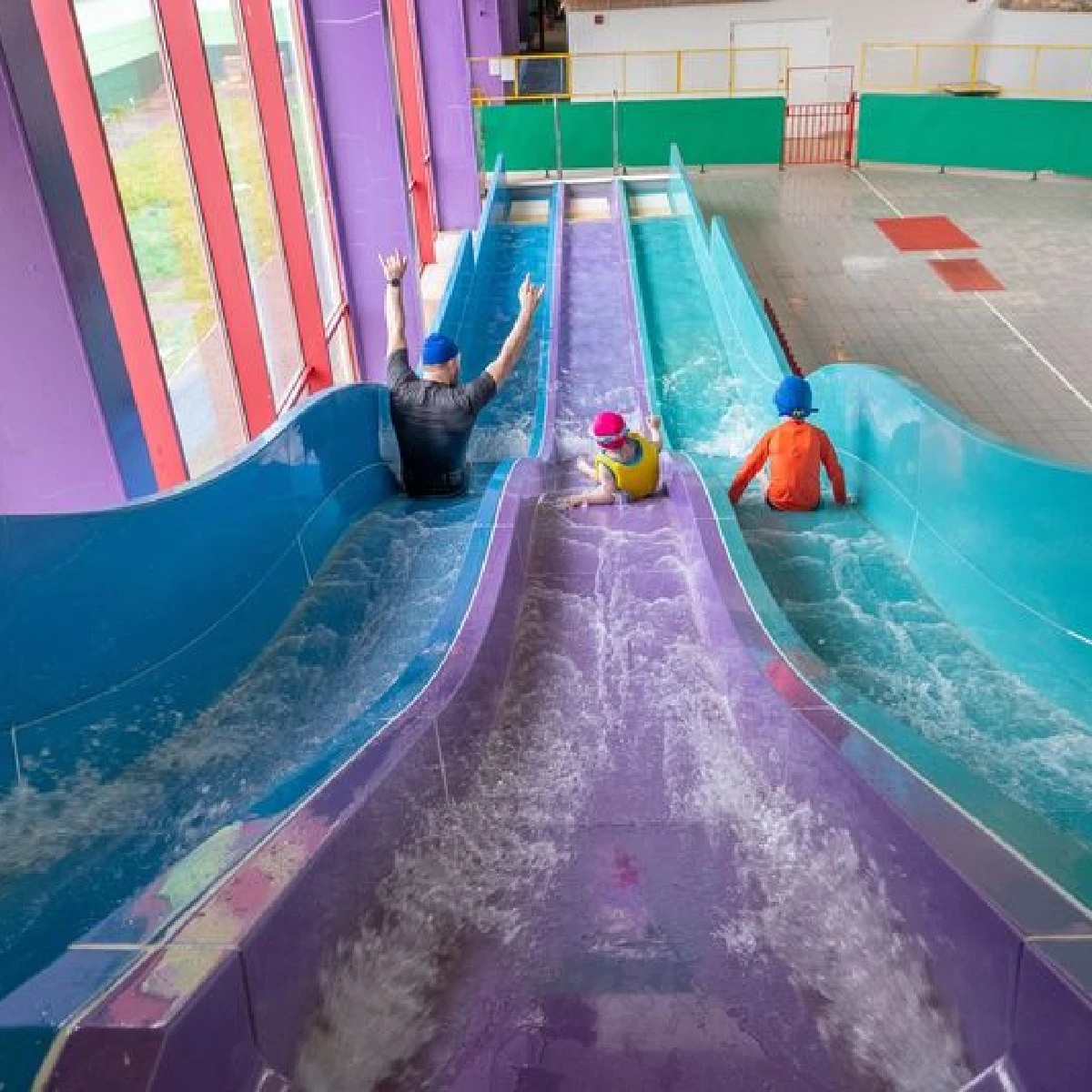 Three people, one adult and two children, slide down colorful water slides indoors. The adult raises both hands in excitement on the leftmost slide, while the children, each on their own slide, descend parallel to the adult. All wear blue swim caps.