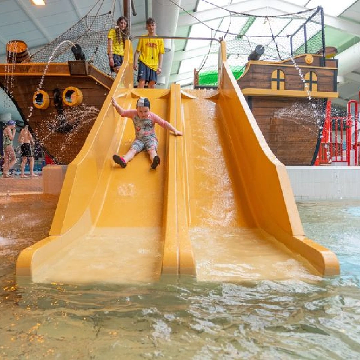 A young child wearing a swimming cap and pajamas slides down a yellow double slide at an indoor water park. In the background, two children in yellow shirts stand beside a pirate ship-themed structure with rope nets. Water splashes at the bottom of the slide.