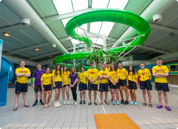 A group of young people wearing yellow shirts and two individuals in purple shirts stand together in front of a large green water slide inside a brightly lit indoor aquatic center. They are posed in a line, smiling at the camera.