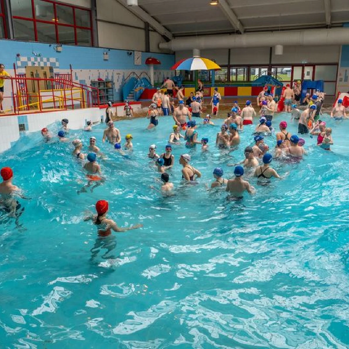 A crowded indoor swimming pool filled with people, mainly children, wearing swim caps. Lifeguards are present, and colorful pool toys are scattered around. The background shows walls with aquatic-themed decorations and large windows letting in natural light.