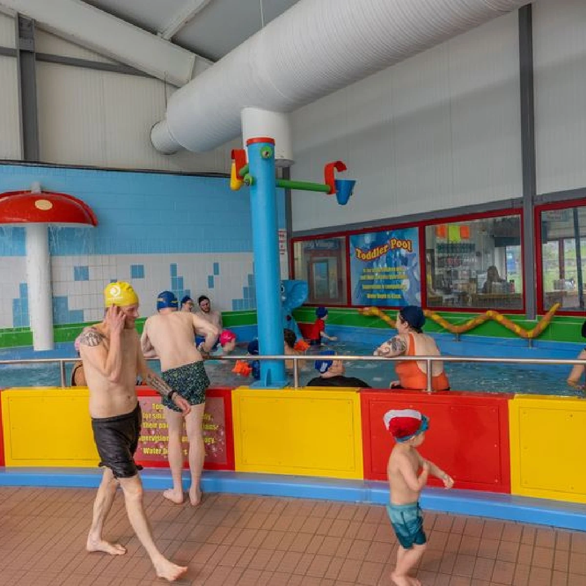 Children and adults in swimwear enjoy a brightly colored indoor pool area. A child in a yellow swim cap and a man in green trunks stand by the pool. In the background, other individuals are in the water near a play structure with pouring cups.