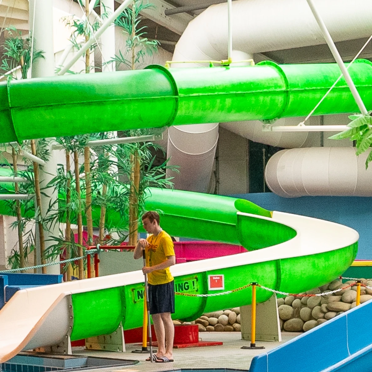 A person in a yellow shirt stands at the entrance of a large, twisting green water slide inside an indoor water park. The area is surrounded by green artificial plants and various white pipes and tubes. A "No Diving" sign is visible near the slide.