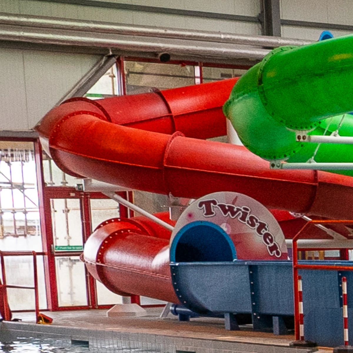 Colorful water slides inside an indoor swimming facility. The slides are red, green, and blue, twisting and turning above the pool area. The entrance to the red slide is labeled "Twister." The slide's structure is supported by metal beams.