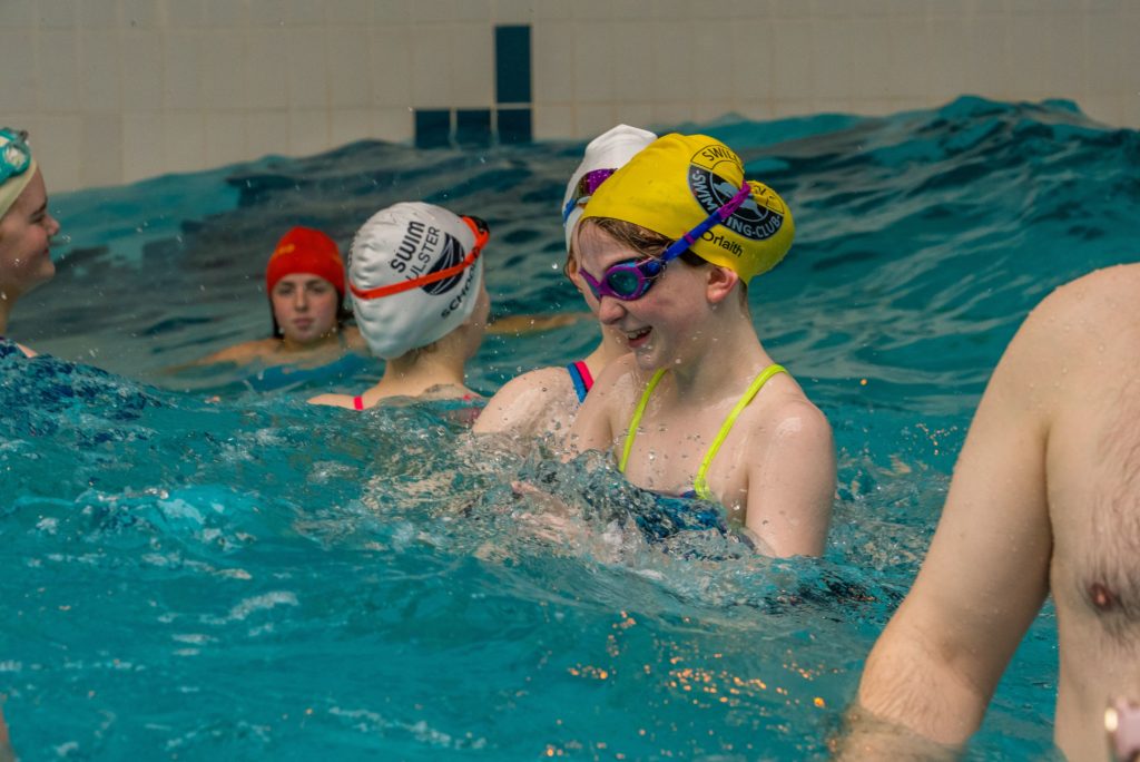 A group of swimmers in a pool, wearing colorful swim caps and goggles. One swimmer in the foreground is smiling and splashing water. Others are visible in the background, engaged in conversation and swimming. The pool tiles are visible in the background.