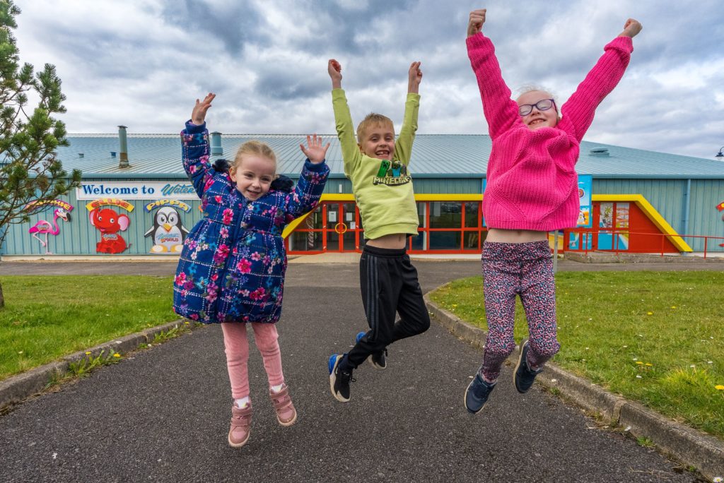 Three children jump joyfully in front of a colorful building with cartoon characters and a "Welcome to..." sign. The child on the left wears a blue floral coat, the middle child wears a green hoodie, and the child on the right wears a pink sweater and patterned pants.