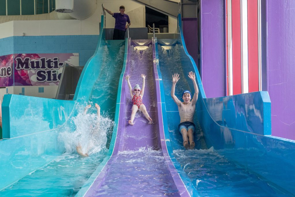 Children slide down three adjacent water slides at an indoor water park. One child in the middle is in a red swimsuit and goggles, while the child on the left splashes into the water, and the child on the right raises their arms excitedly.