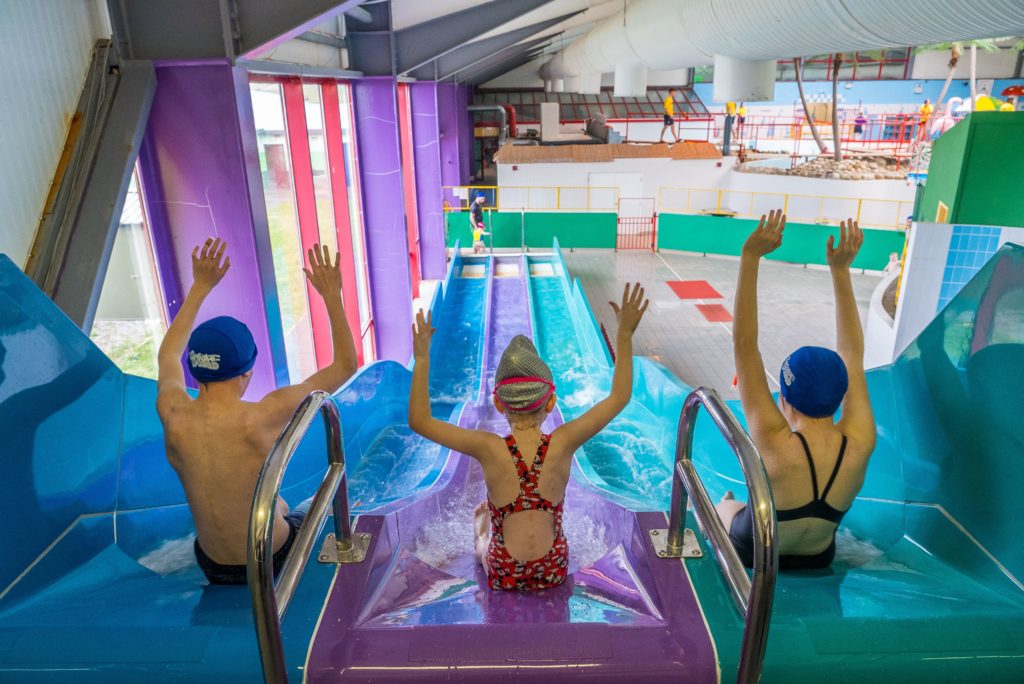 Three children in swimsuits sit at the top of a colorful water slide with their arms raised excitedly, ready to slide down. The indoor water park has various water activities visible in the background, including a pool and play structures.