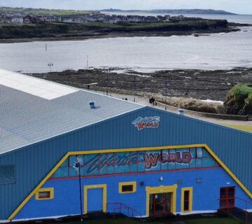 A large blue and yellow building with a sign that reads "Water World" is situated near a rocky shoreline and a body of water. The building features a green roof and several windows. In the background, there are more buildings and a distant landscape with a coastline.